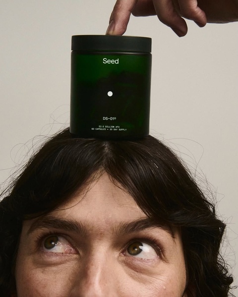 A close-up, high-contrast shot of a human head with a green probiotic jar balanced on top, held by a hand.